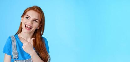 Sassy good-looking dreamy redhead female student contemplate cool performance, tilt head joyfully pointing look left, discuss holiday plans, curiously asking question, posing blue background photo