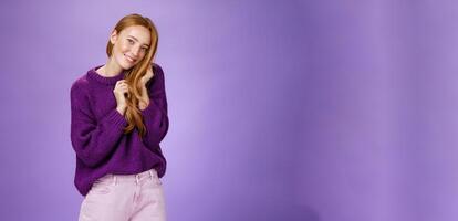 Tender and flirty feminine redhead woman playing with hair tilting head to shoulder and smiling cute and sensual as if making gazes of admiration, being coquettish over purple background photo