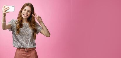 Sociable attractive glamour young caucasian woman taking selfie show victory peace sign smiling broadly raise smartphone look grinning mobile phone display, calling friends, pink background photo