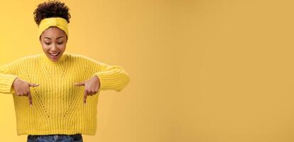 Impressed enthusiastic smiling fascianted african-american woman in sweater headband look pointing down index fingers gasping astonished fascinated cool thrilling promo, standing yellow background photo