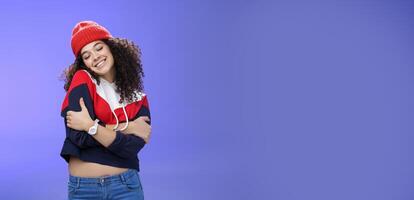 Sweet tender and sensual curly-haired woman in red stylish beanie hugging herself with closed eyes and nostalgic smile leaning on shoulder feeling warmth and tenderness as recalling loving memories photo