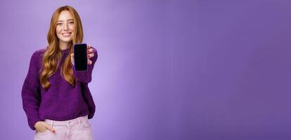 Girl shows smartphone screen at camera to ask opinion of friend smiling broadly with optimistic and joyful expression holding hand in pocket promoting mobile phone or app over purple background photo