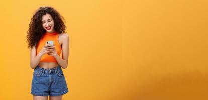 Girl spending time in internet texting friend messages via smartphone laughing while looking at device screen standing happy and upbeat over orange background in cropped top and denim shorts photo