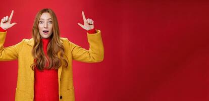 Surprised redhead girl in yellow coat open mouth impressed and thrilled as raising hands and pointing up telling about cool promotion with excited expression against red background photo