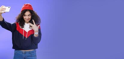 Cool and stylish good-looking caucasian female with curly hair in trendy red beanie and sweatshirt looking from under forehead awesome and swah taking selfie with smartphone holded in extended arm photo