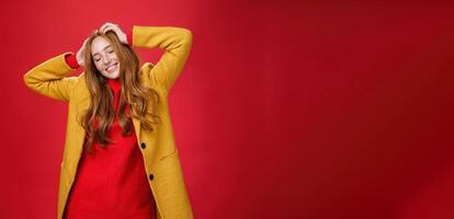 Carefree happy ginger girl carrying away with music and good vibes holding hands on head close eyes and smiling delighted and relaxed having fun, dancing against red background in yellow coat photo