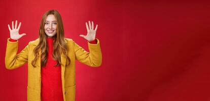 Cute and tender good-looking friendly redhead woman with blue eyes and freckles in stylish yellow coat showing number ten or ordering dozen of things as smiling happily at camera over red background photo