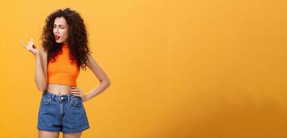 Portrait of moody arrogant and displeased european female with curly hairstyle and cropped top showing small and tiny object with fingers looking at little thing with disdain over orange background photo