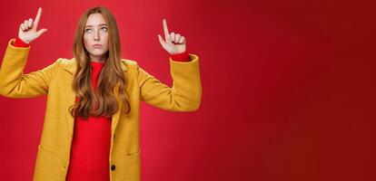 Sad and sorrow cute redhead female in yellow coat raising hands up displeased with regret looking at upper left corner apathical as being disappointed with rain or bad weather over red wall photo