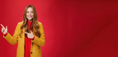 Cheerful friendly-looking and energized helpful redhead girl in yellow coat showing way pointing at upper left corner and smiling broadly with satisfied grin posing delighted over red background photo