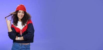 Stylish and snobbish arrogant curly-haired woman in warm beanie rolling curl on finger smirking and looking with contempt at camera, scorning person as being too cool over blue background photo