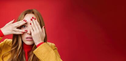 Stunned and shocked redhead woman cannot believe what terrible thing she sees covering face with palms open mouth from shook and peeking through fingers anxious and scared over red background photo
