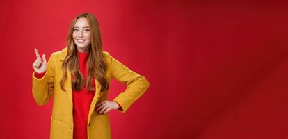 Charming young female in yellow coat holding hand on waist and pointing at upper left corner, smiling broadly as showing or promoting interesting proposal over red background, in good mood photo