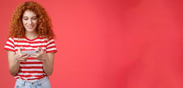 Redhead girl fool around waiting queue dentist playing awesome smartphone game hold phone horizontal tap cellphone screen look telephone display smiling delighted entertained standing red background photo