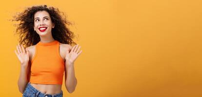 Carefree and happy stylish urban female with nose ring and curly hairstyle raising palms in clueless and indifferent gesture looking up in sun joyfully with delighted cute smile over orange background photo