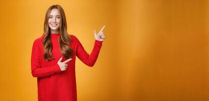 Friendly-looking joyfuly and energized redhead female in red sweater pointing at upper right corner promoting advertisement with broad delighted and tender smile posing against orange background photo