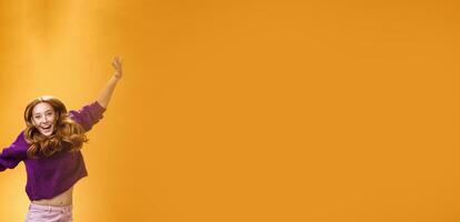 Expressive and enthusiastic carefree friendly joyful redhead woman in purple sweater and pants jumping joyfully with raised hands smiling yelling from happiness and joy against orange background photo