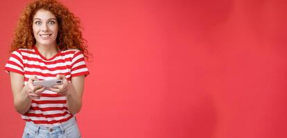 Excited happy cute redhead curly-haired girlfriend eager start learn how play boyfriend game look amused cheerful camera smiling joyful hold horizontal smartphone load app red background photo