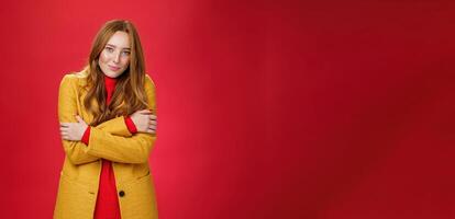 Romantic good-looking tender and cute ginger girlfriend with freckles hugging herself and looking with sensual gentle smile at camera getting cold or chilly waiting outside over red background photo