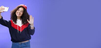 Studio shto of stylish cute female vlogger with curly hair in winter red hat smiling and waving hello at smartphone camera as recording , making interesting content to post online over blue wall photo