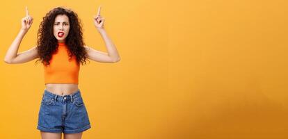 Indoor shot of displeased female client with curly stylish hairstyle red lipstick in cool cropped top pointing up demanding explanation with confused and dissatisfied expression over orange background photo