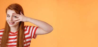 Charismatic lovely redhead lucky female, smiling cute, peek camera tender coquettish gaze, hold palm face, look through fingers, stand orange background optimistic, cheerful mood photo
