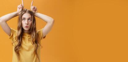GIrl going crazy from boredom making freaky funny faces fooling around squinting holding index fingers on head like horns sticking out tongue and folding lips aping over orange background photo
