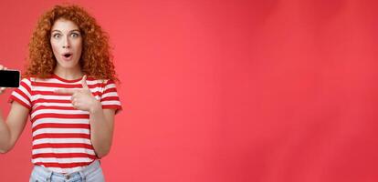 Amused fascinated cute redhead curly girl adore playing games hold smartphone horizontal show thumb up open mouth gasping amazed happy cheerfully present own score favorite app red background photo