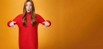 Wow what awesome promotion. Portrait of impressed and surprised cute wondered redhead woman in stylish warm knitted dress folding lips in amazement pointing down and looking at camera photo