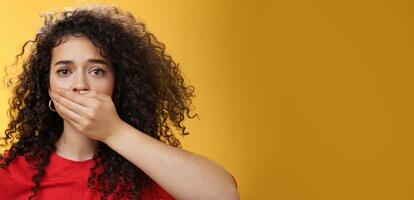Close-up shot of concerned and insecure troubled woman keep silent scare of telling anyone her problem covering mouth with palm not to scream or slip word looking sad and sorrow over yellow background photo