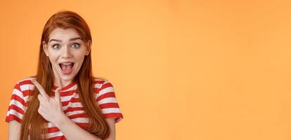Excited female redhead hear impressive sale offer pointing upper left corner, drop jaw astonished stare camera full disbelief surprise, discuss interesting promotion, stand orange background photo