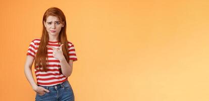 Upset uneasy cute ginger gitl long natural red hair, frowning smirking hesitant, complain, looking suspicious and doubtful point left, not sure if product really good, feel unsure, orange background photo