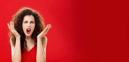 Moody displeased and angry rich woman being dissatisfied with awful chip gift raising palms in frustration and clueless gesture arguing, shouting in outrage standing in red elegant dress photo