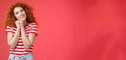 Tender feminine caring redhead curly-haired female romantic gaze camera tilt head clench palms near head silly lovely look smiling delighted cheerful dreamy sighing look with sympathy photo