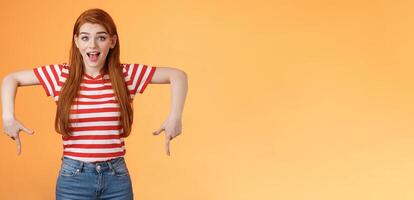 Enthusiastic lively cute redhead woman introduce incredible offer, pointing down, bottom copy space, smiling amazed, look camera, explain you awesome opportunity, feel excitement, orange background photo