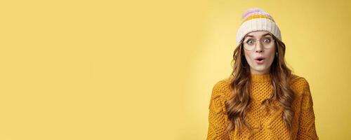 Surprised amazed charismatic good-looking young female student widen eyes wondered say wow amazed standing shocked impressed yellow background, wearing nerd glasses hat sweater photo