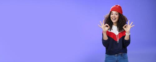 Got under control. Portrait of happy charming smiling curly-haired female in warm winter hat and sweatshirt smiling broadly and showing okay or excellent gesture as approving, liking cool movements photo