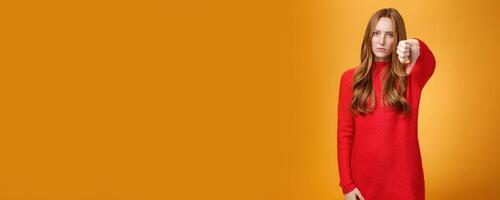 Disappointed upset and serious-looking attractive 20s redhead woman in red knitted dress frowning in dislike looking with judgement, showing thumb down in aversion and dissatisfaction over orange wall photo