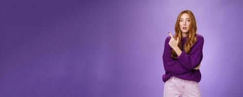 Astonished excited redhead woman open mouth and raise eyebrows questioned as seing shocking thing pointing at upper left corner, asking question impressed and surprised over purple wall photo