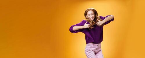Optimistic and joyful bright ginger girl jumping happily and cheerful having fun showing thumbs up in like and approval giving positive feedback of super cool idea or concept over orange wall photo