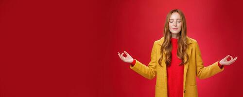Girl keep calm releasing stress with meditation, posing in yellow coat, close eyes and looking relieved as extending hands sideways with mudra gesture, doing yoga against red background in lotus pose photo