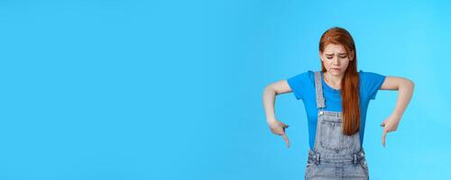 Moody upset cute redhead woman feel uneasy, frowning look pointing up jealousy and regret, cannot afford awesome new product, express sadness and sorrow, stand blue background gloomy photo
