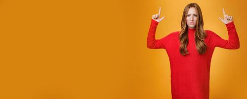 Intense disappointed and distressed attractive young 20s redhead female in red warm sweater frowning upset with dismay raising hands pointing up dissatisfied against orange background photo