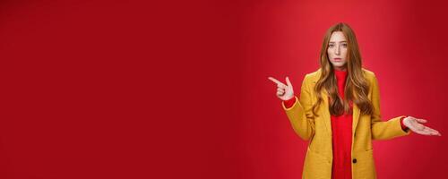Frustrated and confused young cute female with ginger hair in yellow coat shrugging as pointing questioned left waiting explanation as asking question clueless and puzzled over red background photo