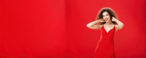 Stylish gentle caucasian girl with curly hairstyle and evening make-up touching strands of hair checking haircut and smiling broadly at camera posing in new elegant red dress over studio background photo