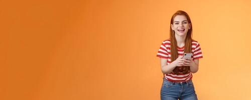 Carefree attractive woman red long hair laughing amused, having fun, chatting friend, hold smartphone, look camera sincere rejoice smile, texting joke, found funny meme internet, orange background photo