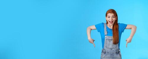 Amused wondered excited cute 20s european woman, pointing down, bottom copy space, look amazed admiration camera, describe amazing product, recommend site, stand blue background photo