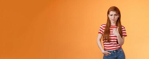 Upset uneasy cute ginger gitl long natural red hair, frowning smirking hesitant, complain, looking suspicious and doubtful point left, not sure if product really good, feel unsure, orange background photo