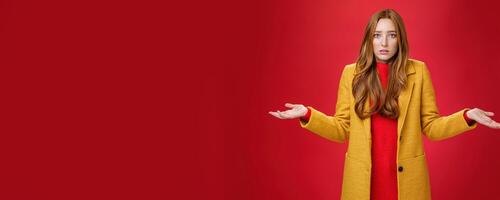 Upset and confused redhead girlfriend cannot understand what happened shrugging with spread hands sideways and clueless grimace, being unaware why, posing concerned over red wall photo