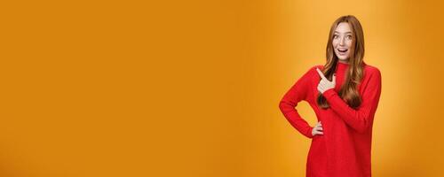 Energized optimistic and happy friendly-looking caucasian ginger girl in red sweater pointing at upper left corner with enthusiastic excited and joyful smile posing delighted against orange background photo
